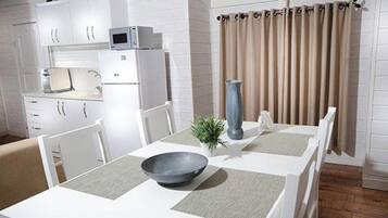 Een kleine keuken met witte kasten, een magnetron en een koelkast. Er staat een eettafel met een kom en een vaas erop.