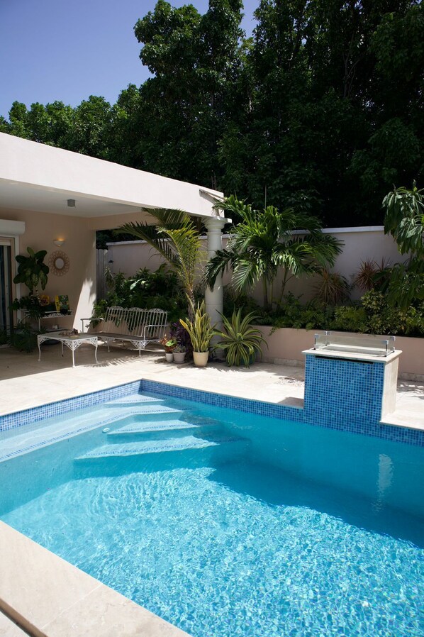 A swimming pool with a tiled edge, surrounded by potted plants and a white outdoor seating area.
