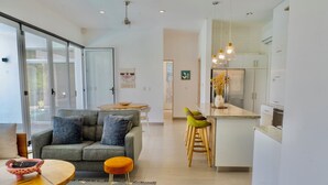 Eine moderne Küche mit weißen Schränken, einer zentralen Kochinsel und einem Essbereich mit grauem Sofa und orangefarbenem Hocker.