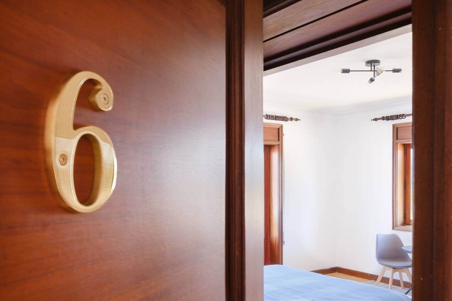 A hotel room with a wooden door marked with the number 6, a bed, a chair, and a ceiling fan.