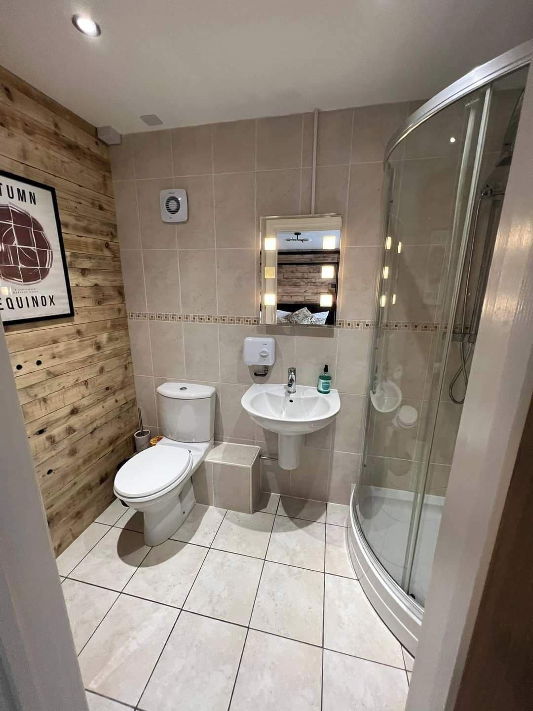 Kylpyhuone, jossa on puinen seinä, wc-istuin, pesuallas, valaistu peili ja lasinen suihkuseinä.