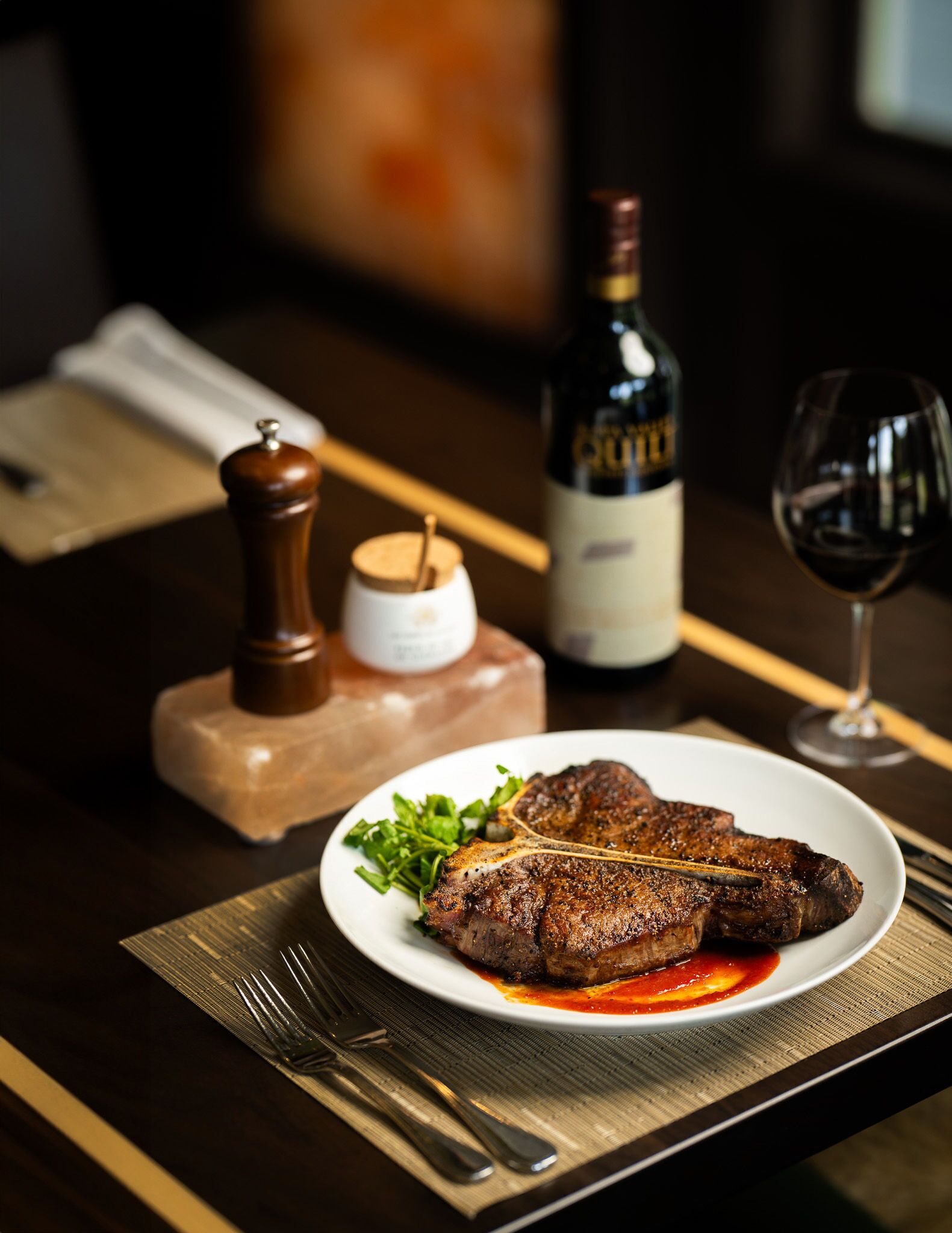 A plate of steak with a side of greens, a bottle of wine, and a glass of wine on a table.