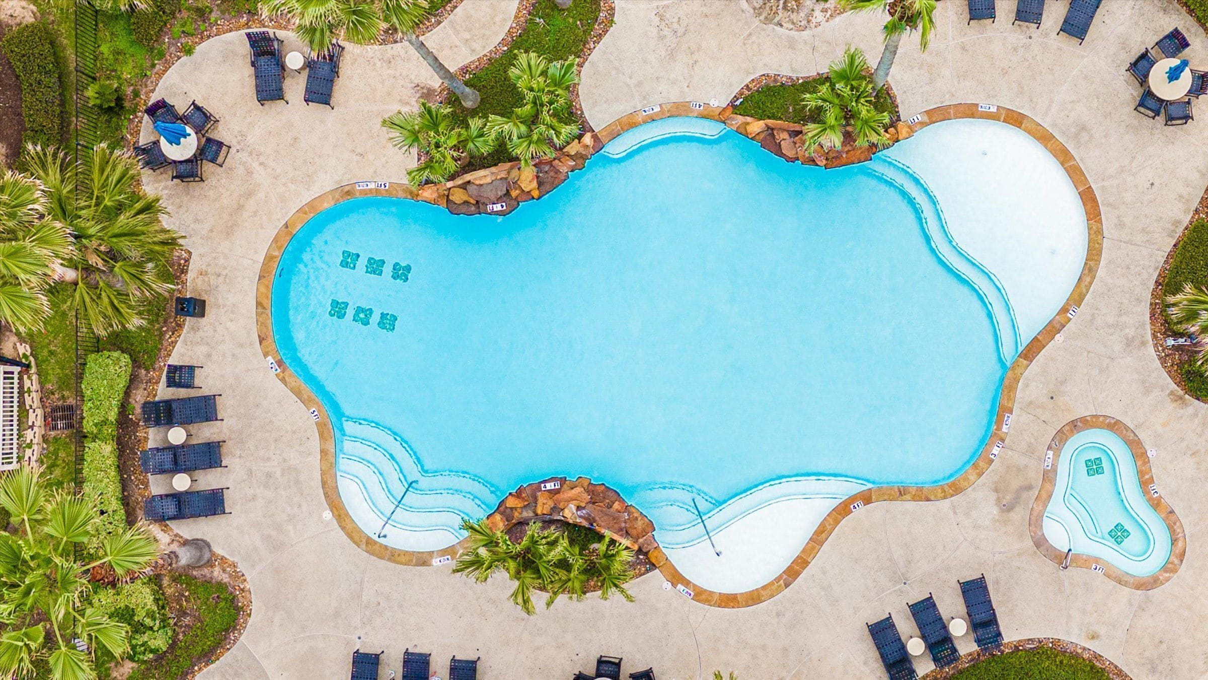 An aerial view of a swimming pool with surrounding lounge areas, palm trees, and a stone border.