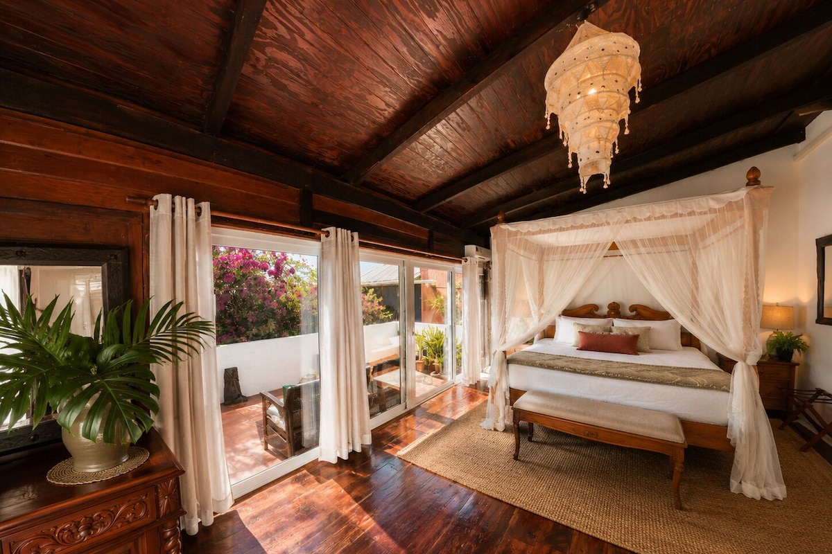 Ein Schlafzimmer mit Baldachinbett, Holzboden, einem großen Fenster mit Vorhängen und einer Topfpflanze.