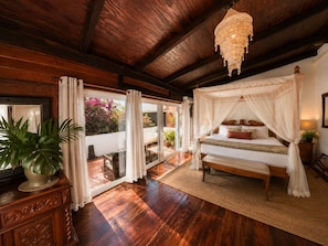 A bedroom with a canopy bed, wooden flooring, a large window with curtains, and a potted plant. - Crystals Villa Hotel (Soufriere)