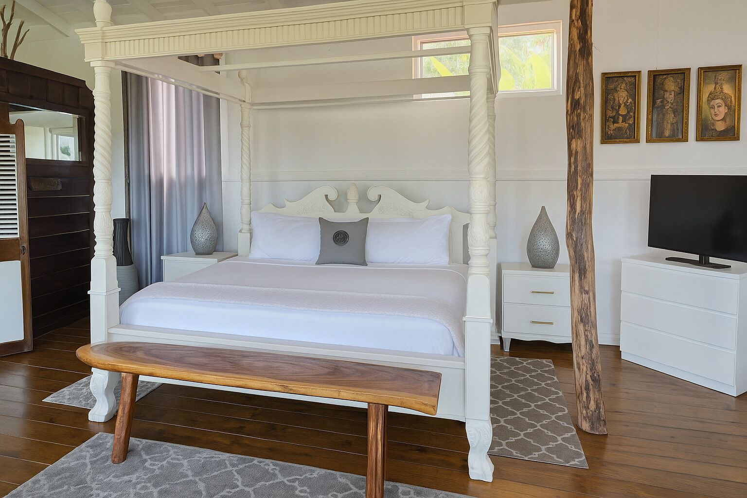 Un lit à baldaquin avec un banc en bois devant, des tables de chevet blanches, un téléviseur posé sur une commode et des œuvres d’art encadrées accrochées au mur.