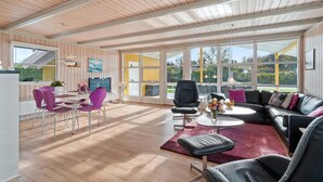 Een ruime woonkamer met een houten plafond, grote ramen, een zwarte leren bank, paarse stoelen, en een eethoek met een televisie en kaarsen.
