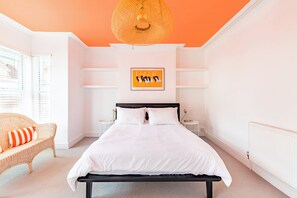 A bedroom with a bed, a wicker chair, a bedside table, a wall-mounted shelf, and a hanging light fixture.