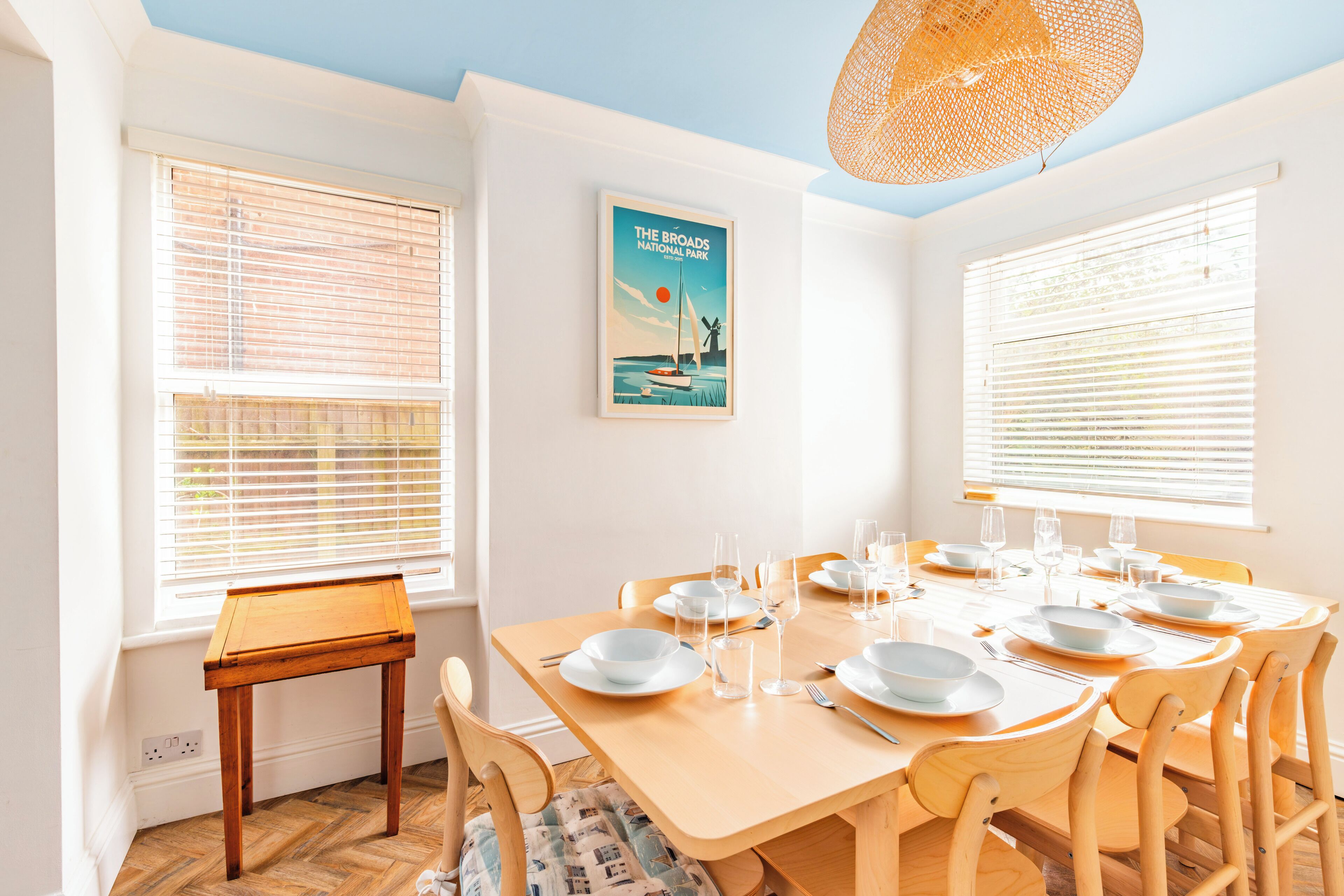 Une salle à manger avec une table en bois dressée pour six personnes, des chaises blanches et une affiche du parc national de The Broads accrochée au mur.