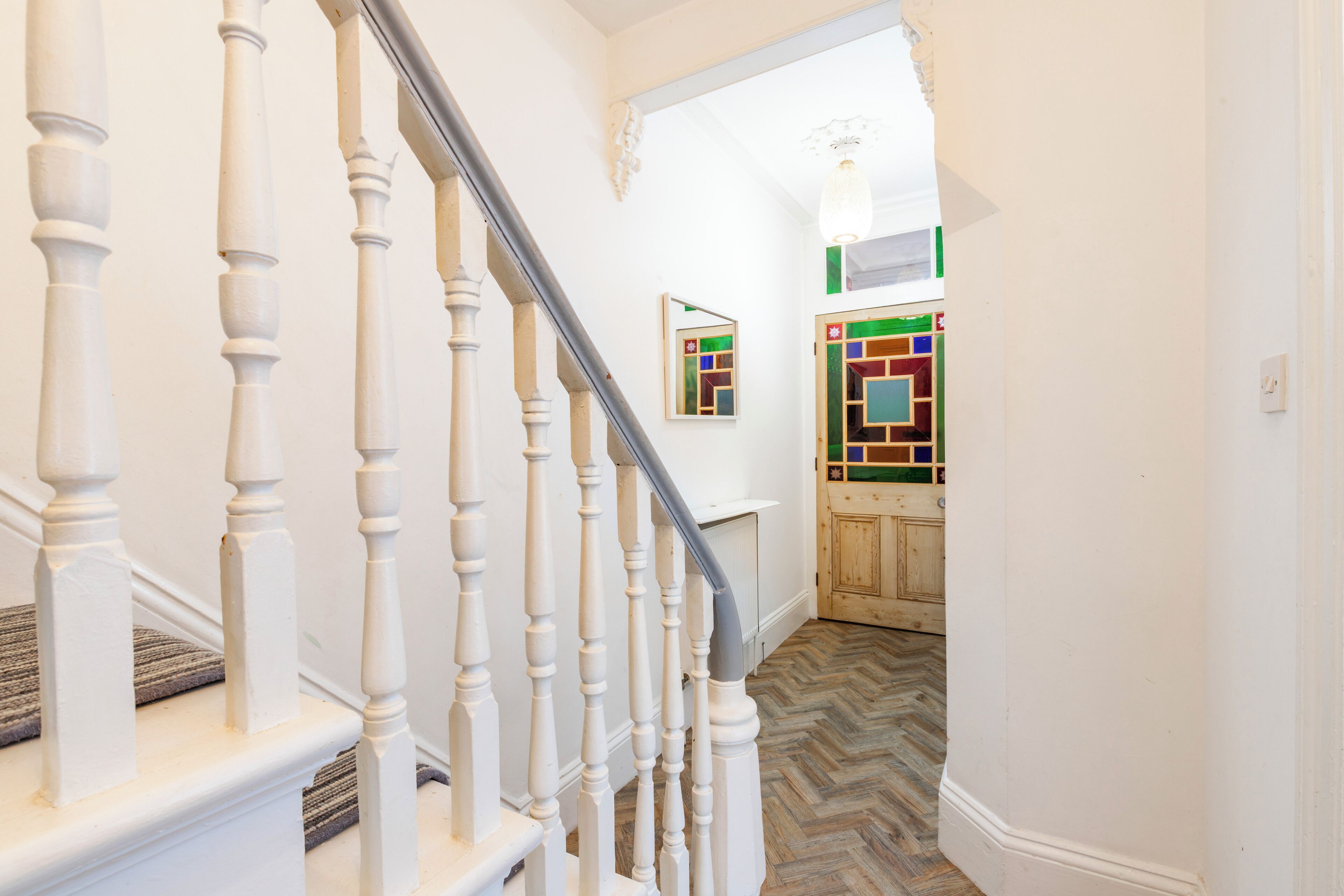 Un escalier avec des balustrades blanches menant à un palier, un parquet à motifs en chevrons et une porte en bois ornée d’un vitrail aux couleurs vives.