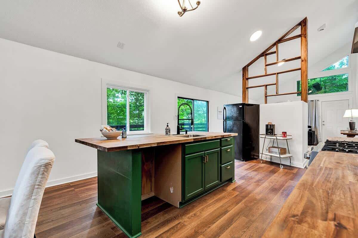 Une cuisine dotée d’un plan de travail en bois, de meubles de rangement verts, d’une cuisinière encastrée et offrant une vue sur la verdure par les fenêtres.