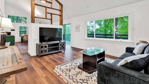 A modern living room with a TV, wooden flooring, and a large window offering a view of greenery.