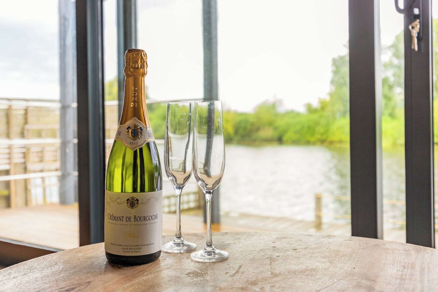 Una botella de champán Cuvée de Bourgogne y dos copas de champán vacías sobre una mesa con vista a un río y vegetación.