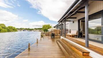 Une terrasse en bois s’étendant au-dessus d’une étendue d’eau, avec un bâtiment moderne doté de grandes fenêtres et d’un espace salon extérieur.
