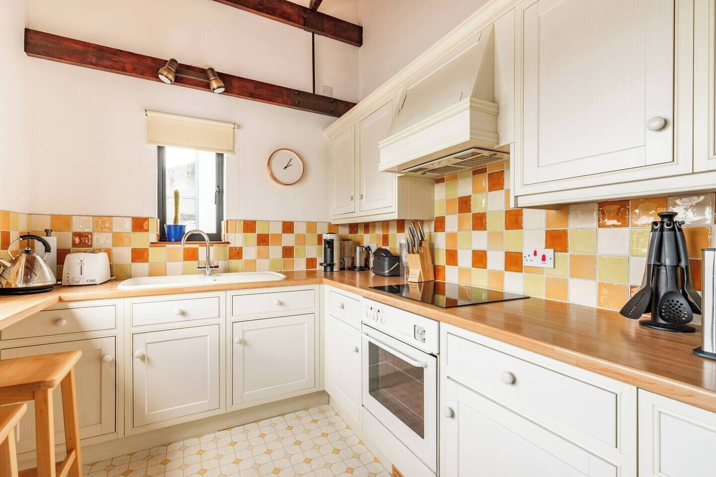 Une cuisine avec des meubles blancs, un plan de travail en bois, un pare-feu en carrelage, une horloge et une fenêtre avec un store.
