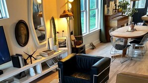 Een moderne woonkamer met een bruine leren bank, een blauwe fauteuil en een glazen salontafel. Er is een boekenplank met boeken en decoratieve objecten, en een groot raam zorgt voor natuurlijk licht.