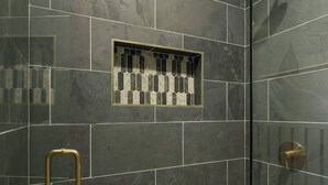 Une douche avec des carreaux foncés, un motif de carreaux hexagonaux en bas et une poignée dorée.