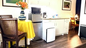 Eine kleine Küche mit Kühlschrank, Mikrowelle und Kaffeemaschine. Ein Esstisch mit Stühlen und eine Blumenvase stehen bereit.