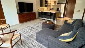 A modern living room with a grey sectional sofa, wooden chairs, a TV, a ceiling fan, and a kitchen area in the background. - Versalles Corner (Puerto Vallarta)