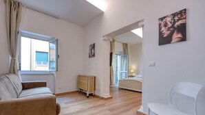 Quarto com piso de madeira, um sofá bege, uma cadeira branca, um armário de madeira e uma cama com mesa de cabeceira.