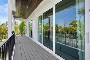 A covered outdoor balcony with a wooden floor, large sliding glass doors, and a view of palm trees and a clear sky.