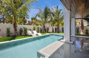 A swimming pool with lounge chairs, surrounded by palm trees and a white fence. - Brand New beach house! Heated pool and spa! (Bradenton Beach)