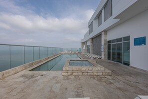 A rooftop pool area with a glass balustrade, lounge chairs, and a clear view of the sky.