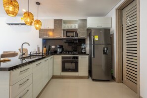 A modern kitchen with a stainless steel refrigerator, a built-in oven, and a microwave. There are hanging pendant lights and a countertop with a coffee maker and a toaster.