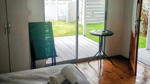 A hotel room with a bed, a green chair, a small table, and a sliding glass door leading to an outdoor area with a wooden deck and a white fence. - Studio 1 Je Nous between the Sea and the Lagoon (Faro de José Ignacio , la Juanita)
