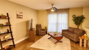 Un salón con piso de madera, un ventilador de techo, un sofá marrón, un sillón marrón, una mesa de centro de madera, una mesita auxiliar, una planta, un cuadro en la pared y una estantería con objetos decorativos.