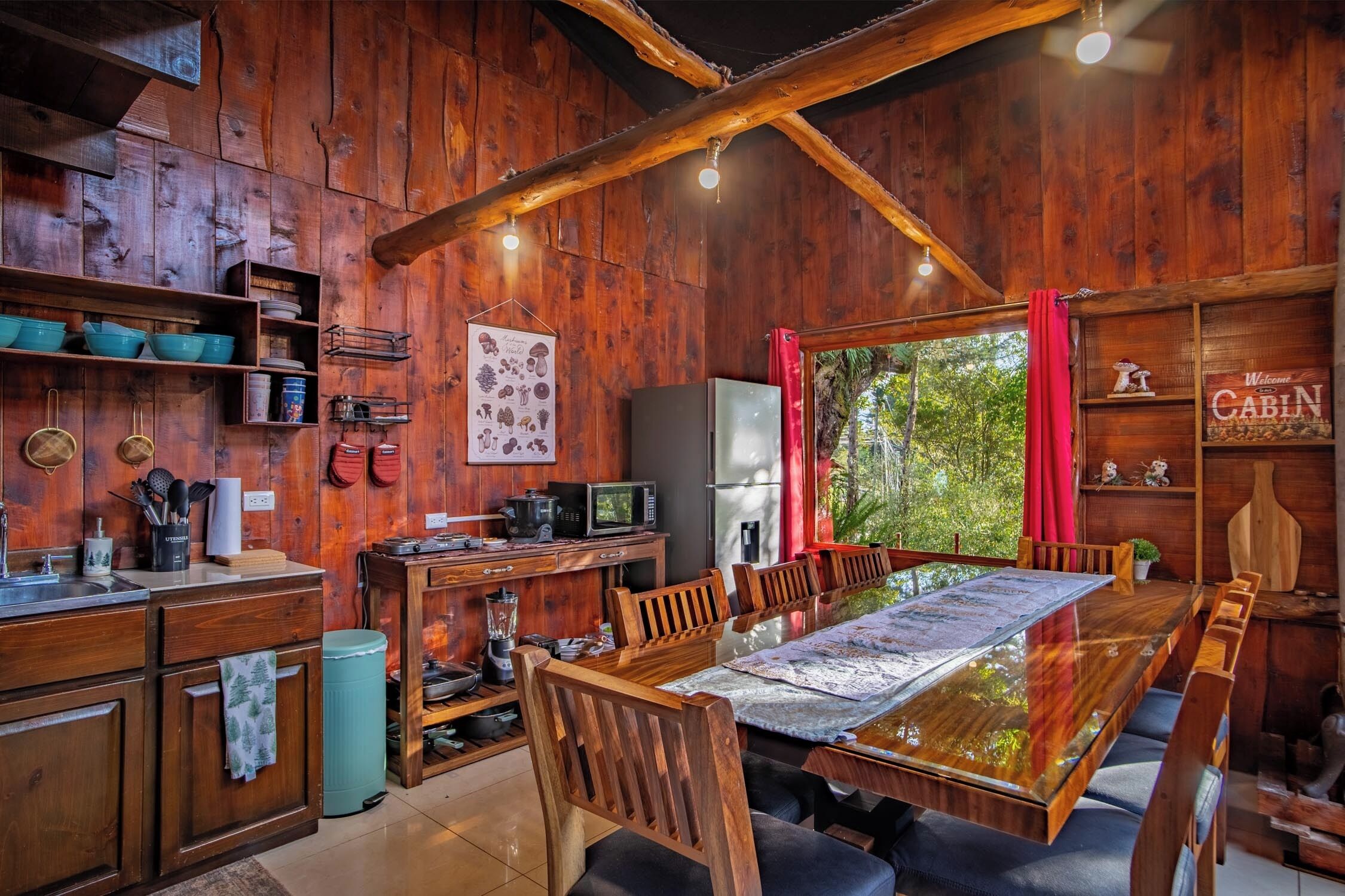 A rustic cabin interior with wooden walls, a dining table, chairs, a kitchen area with a refrigerator, microwave, and various kitchen utensils.