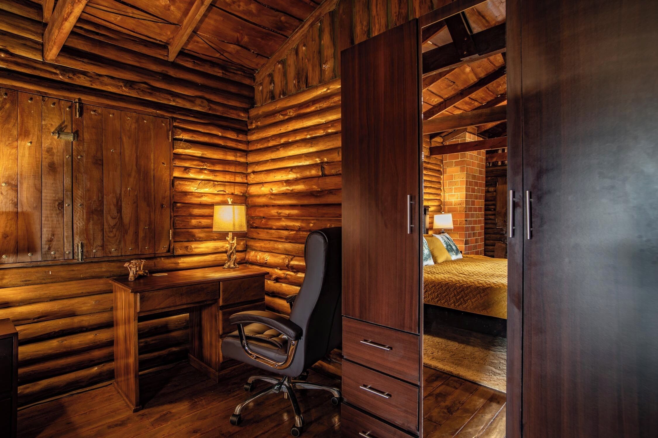 L’intérieur d’une cabane en rondins, avec un bureau en bois, une chaise et un lit, le tout adossé à un mur en briques.
