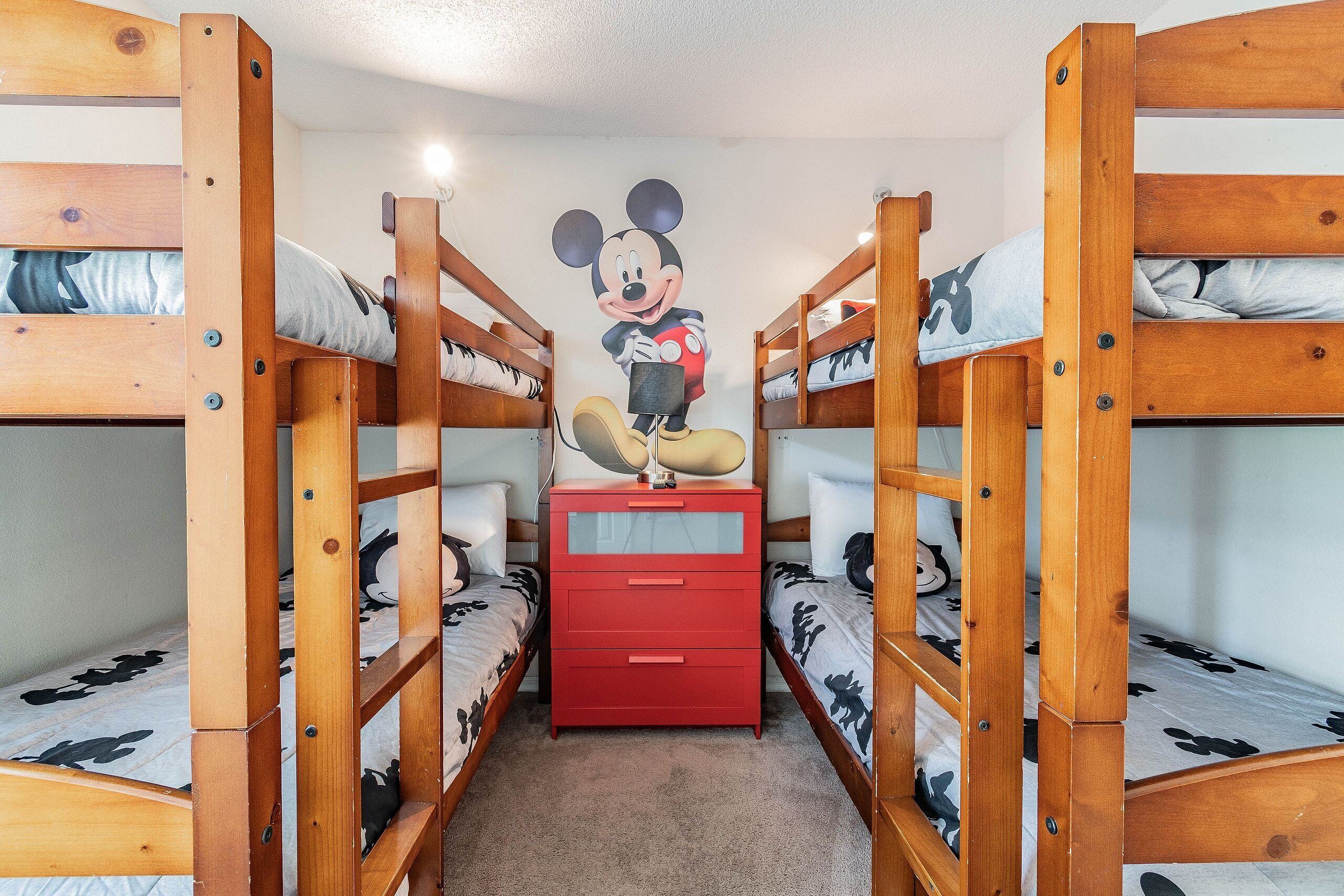 Ein Schlafzimmer mit Etagenbetten, einer Wandmalerei mit Mickey Mouse, einem roten Schminktisch und gemusterten Betttextilien.