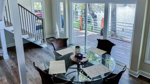 A spacious room with a glass table set for dining, large windows offering a view of a deck and outdoor area, and a staircase leading to another level. - NEW! Lake Front House,Private Dock,Kayaks,Beach (Troutman)