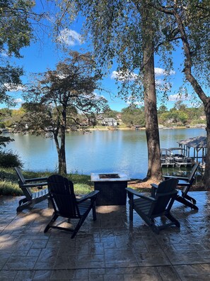 An outdoor seating area with Adirondack chairs and a table, overlooking a lake with boats and houses in the background. - NEW! Lake Front House,Private Dock,Kayaks,Beach (Troutman)