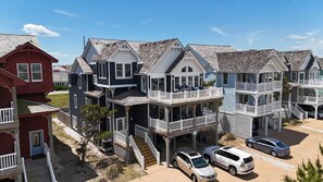 A beachfront property with multiple-story houses featuring balconies, a driveway with parked cars, and a clear sky.