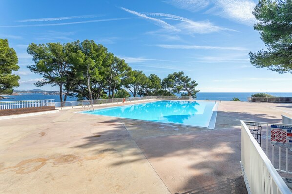 A swimming pool with a view of the sea and mountains, surrounded by trees and a fenced area.