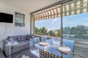 A modern living room with a glass dining table, grey sofa, and a large window with a view of trees and the sky.