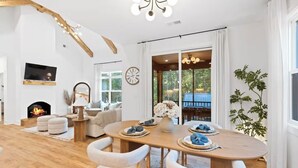 Uma sala de estar espaçosa com lareira, um espaço de jantar com mesa de madeira e cadeiras brancas, e uma grande janela com vista para o verde.