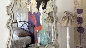 Une chambre à coucher avec un grand miroir décoratif, un lit avec une tête de lit blanche, une table de chevet avec une lampe et un mur recouvert d’un papier peint à motifs floraux.