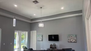 A spacious living room with a black leather sofa, a flat-screen TV mounted on the wall, and two abstract floral paintings. - Cool 3-bedroom house with AC in the Sunshine State (Lehigh Acres)