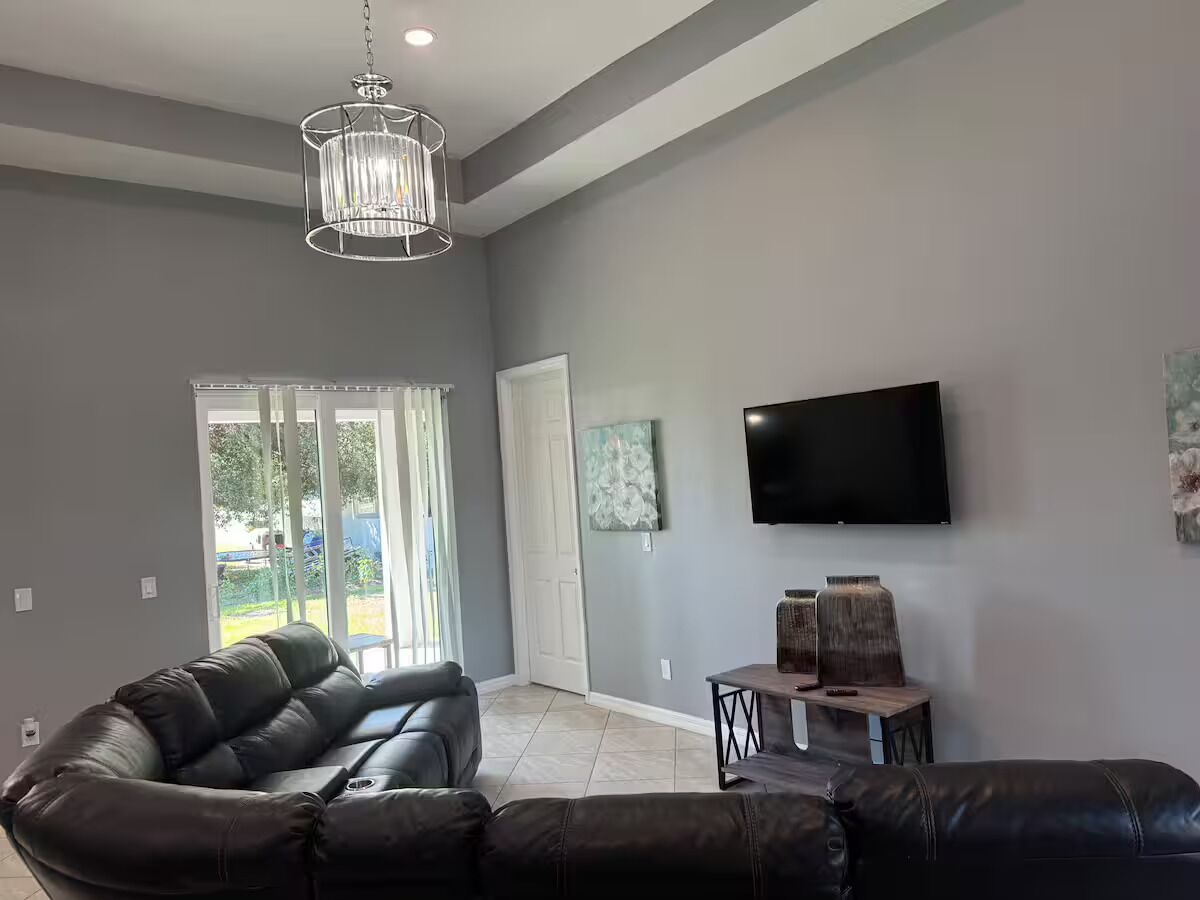A modern living room with a leather sectional sofa, a flat-screen TV mounted on the wall, and a chandelier.