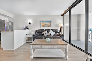 Uma sala de estar moderna com uma mesa de centro de madeira, um sofá de couro preto com almofadas estampadas, uma mesinha lateral branca e um quadro na parede.