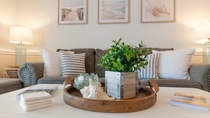Un salon avec un canapé gris, une table basse en bois et des œuvres d’art sur le thème de la plage, encadrées et accrochées au mur.