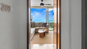 Una sala de estar moderna con un sofá, una mesa de centro y un ventilador de techo. Se ve un balcón con vistas al océano a través de puertas de vidrio.