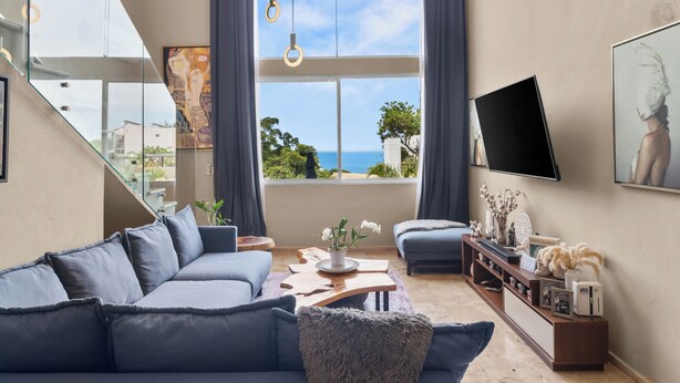 A modern living room with a large sofa, a coffee table, and a TV mounted on the wall. There is a large window with a view of the sea and a painting on the wall.