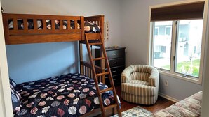 A bedroom with a bunk bed, a chair, a dresser, and a window with a view of buildings. - Beach Block condo, 2 bed, 2.5 bath - Perfect for couples or small families! (Brigantine)