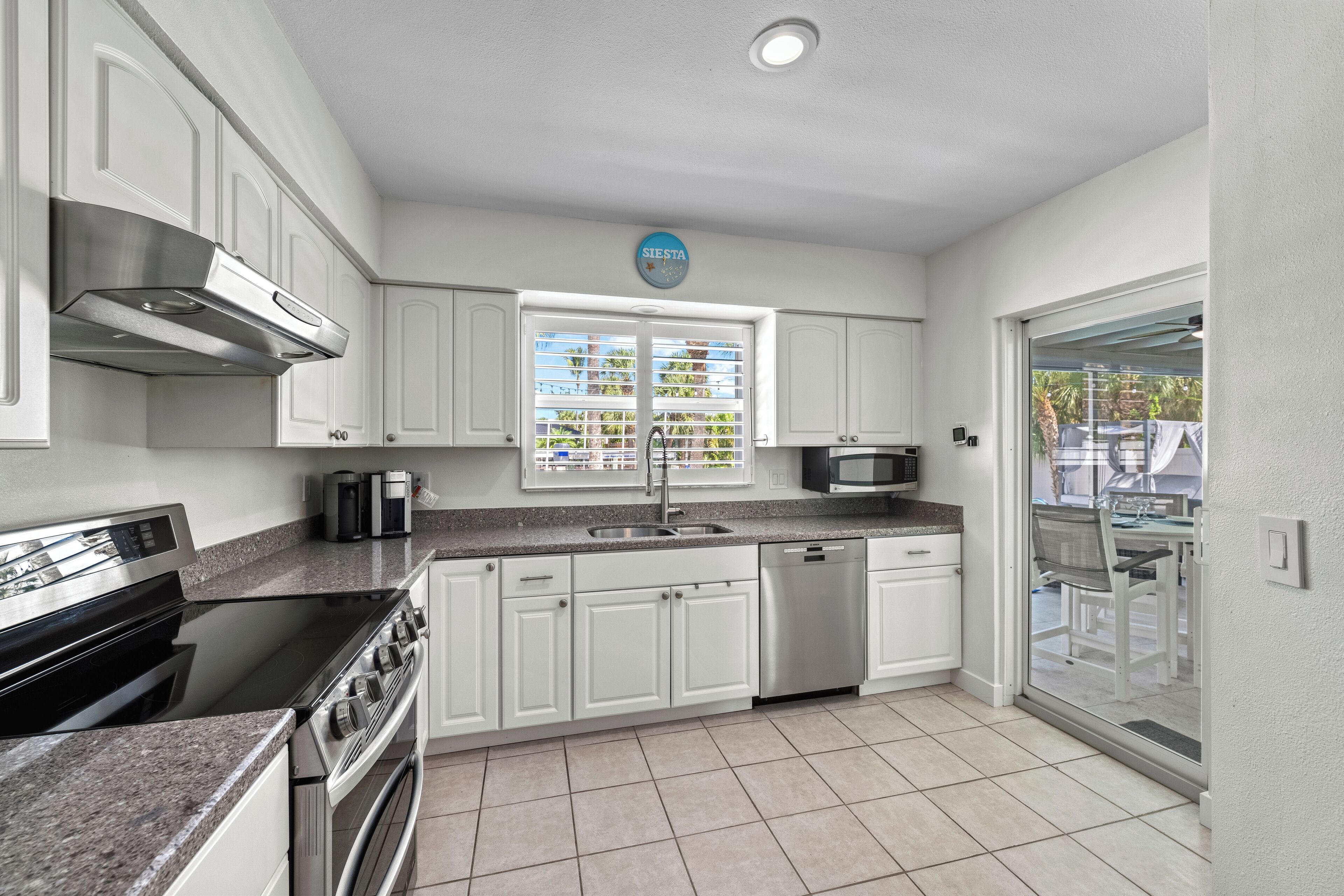 A modern kitchen with white cabinets, stainless steel appliances, a built-in oven, and a window with blinds.