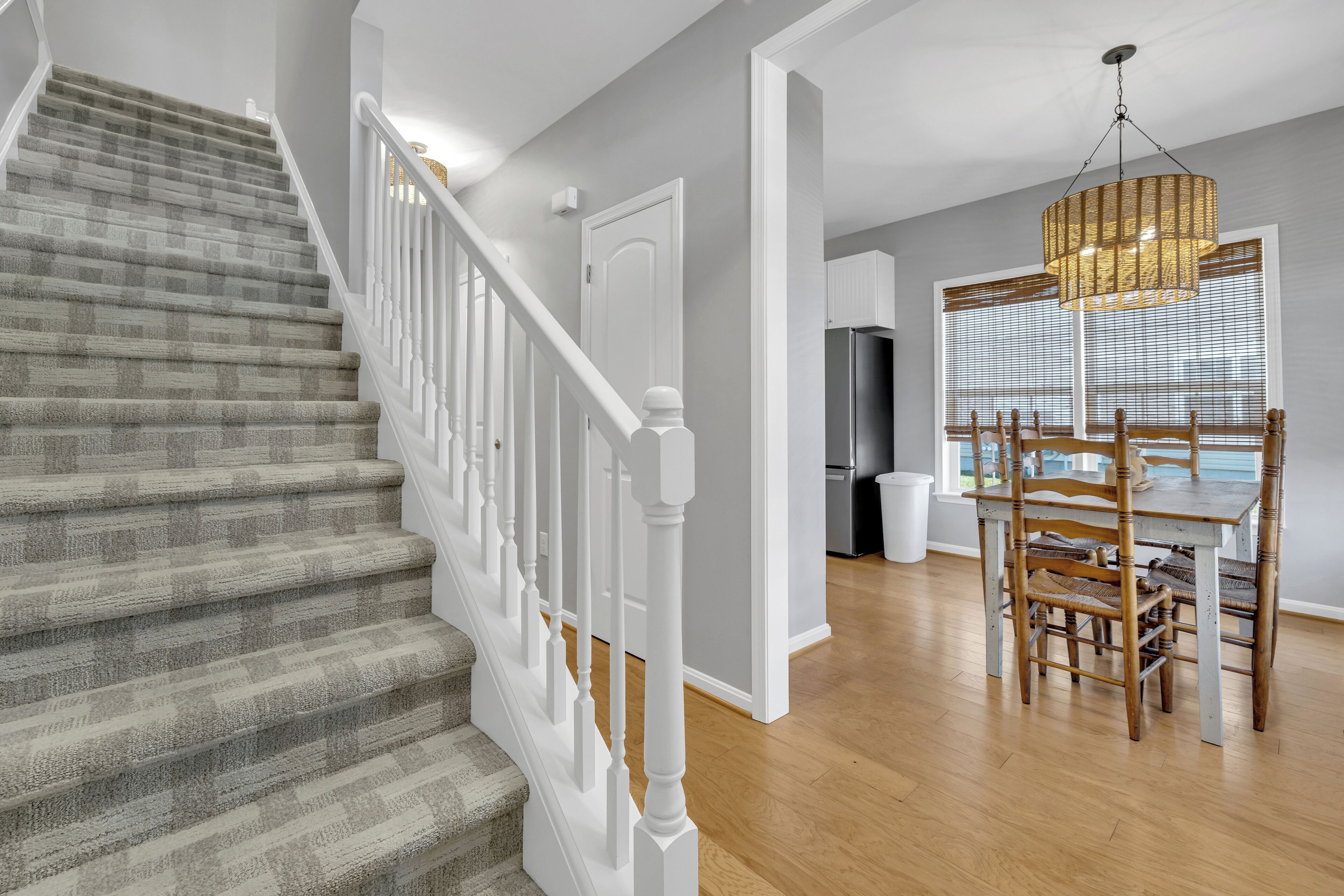 Un escalier avec des rampes blanches, un coin repas avec une table et des chaises en bois, et une cuisine équipée d’un réfrigérateur.