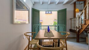 A dining area with a wooden table and chairs, a window with green shutters, and a view of a balcony with plants.